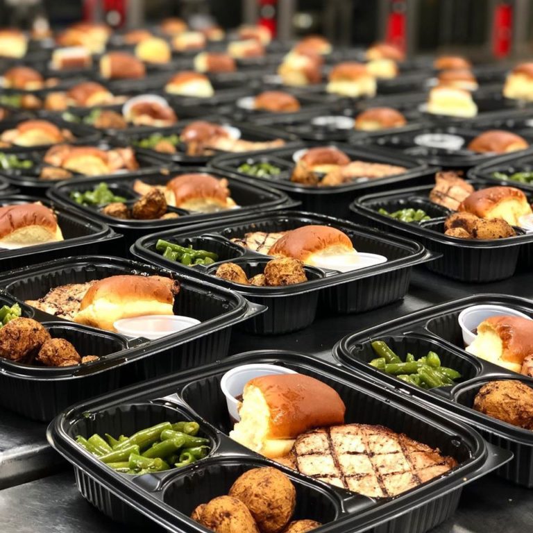 Rows of meal trays with grilled chicken, rolls, green beans, and potatoes neatly arranged on a table.