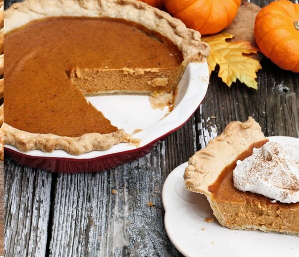 A pecan pie with a slice missing, on a plate with whipped cream, surrounded by small pumpkins and autumn leaves.