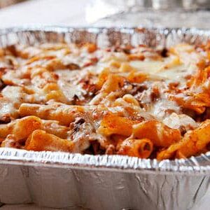 Foil tray of baked rigatoni topped with melted cheese, set on a white surface from Christ the King Family Dinner.