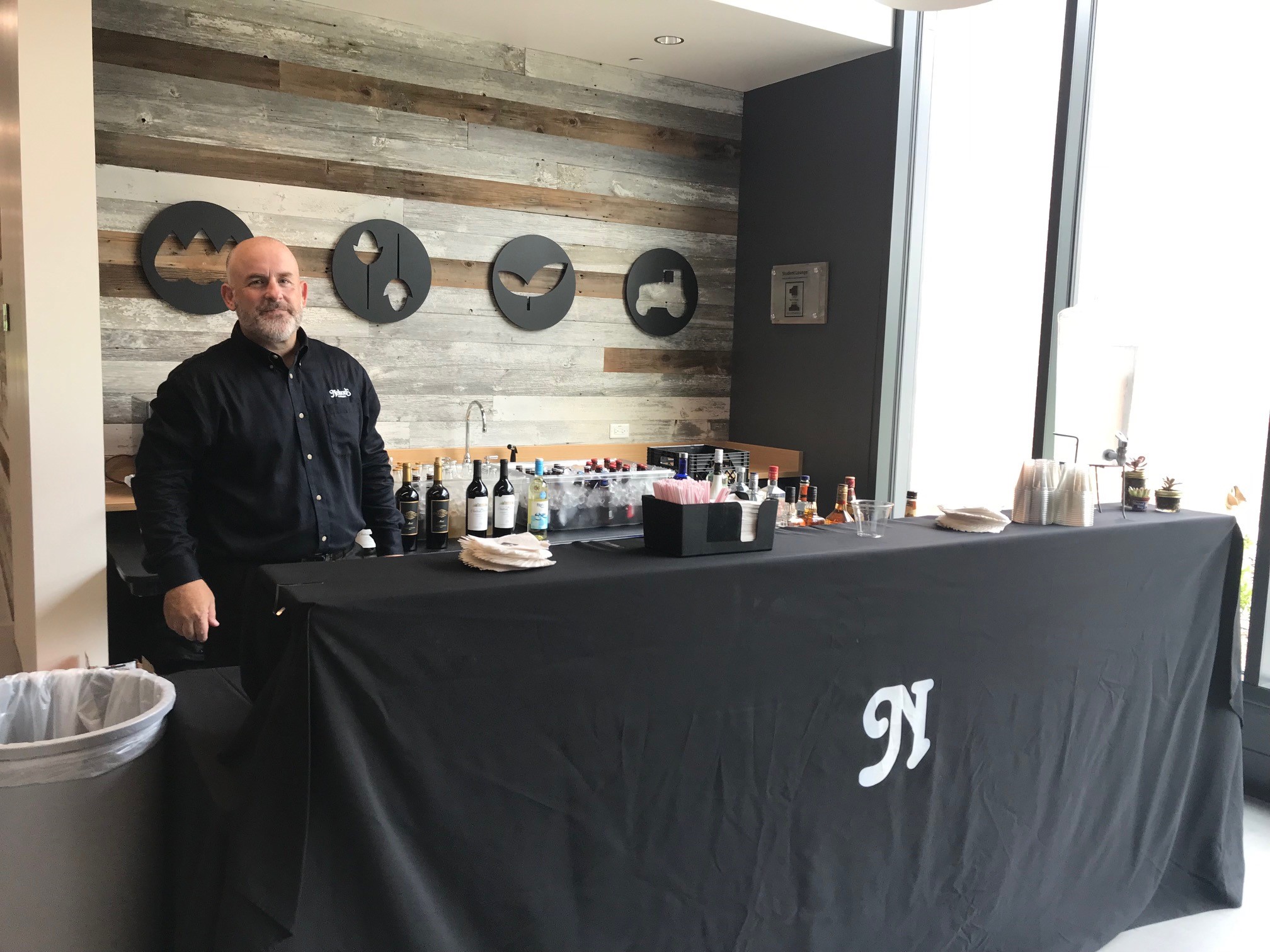 A man in a black shirt stands behind a bar table with drinks and condiments, set in an indoor space with rustic decor.