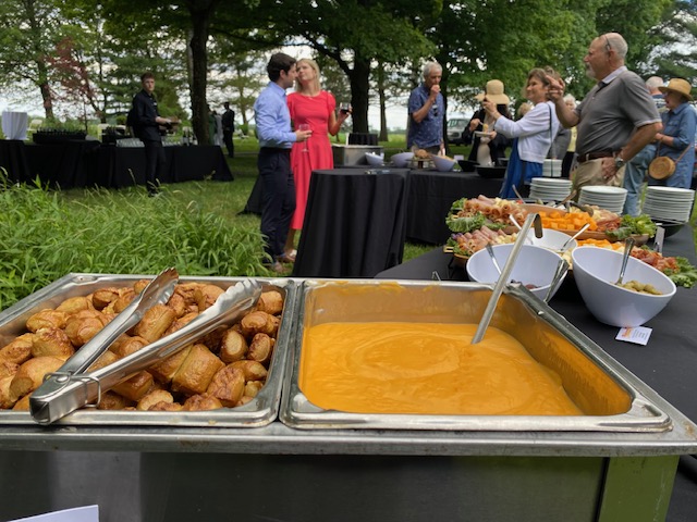 Outdoor buffet featuring pretzel bites, cheese dip, fruit, and olives. People chatting near trees in the background.