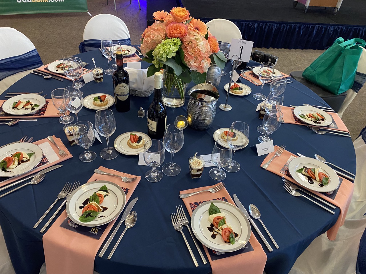 Elegant table with a blue cloth, floral centerpiece, wine, salads on plates, and pink napkins at each setting.