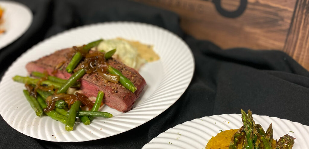 Steak topped with green beans and onions, mashed potatoes, and asparagus on white plates.