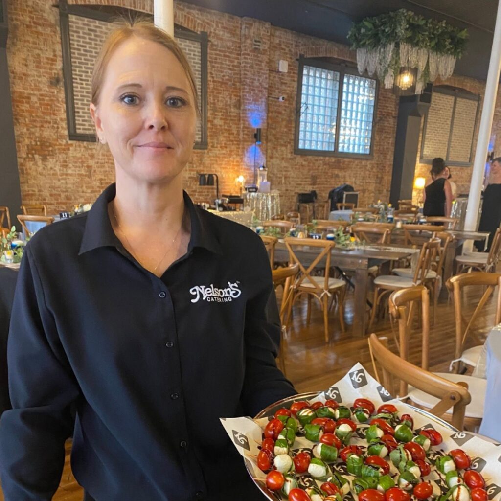A server in a black shirt holds a tray of skewered appetizers in a decorated event space with brick walls and wooden chairs.