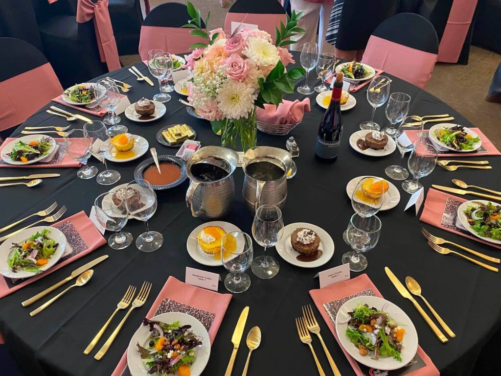 A formal table set for six with salads, desserts, wine, and a pink floral centerpiece on a black tablecloth.